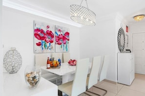 Uma sala de jantar moderna com uma mesa branca, cadeiras e um candelabro. Há quadros abstratos na parede e um vaso decorativo sobre a mesa.