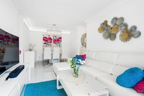 Uma sala de estar moderna com um sofá branco, uma mesa de centro e uma TV. Há peças decorativas nas paredes e um vaso com flores sobre a mesa de centro.