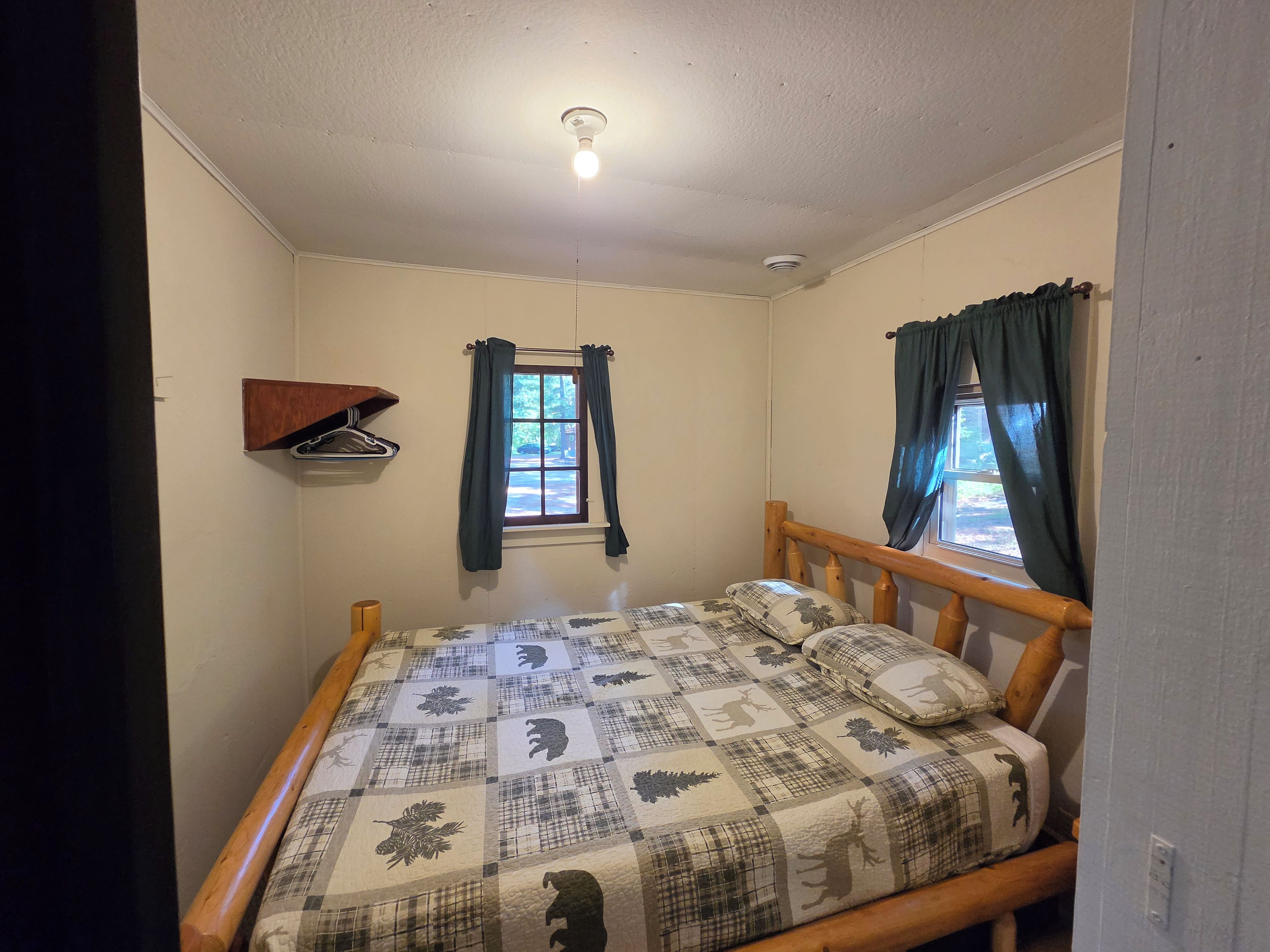 Une petite chambre avec un lit simple, une tête de lit en bois, une fenêtre avec des rideaux, une étagère fixée au mur et un luminaire au plafond.