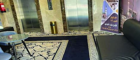 An elevator area with two elevators, a small table with a chair, a blue carpet, and a sofa.
