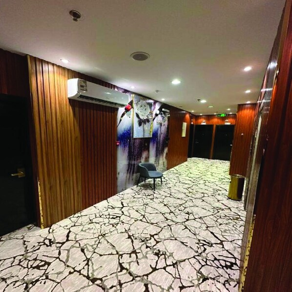 A hotel corridor with a patterned floor, wooden paneling, an air conditioning unit, a chair, and a trash can.