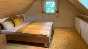 Een slaapkamer met een schuine kap, een bed met een gestreepte deken, een houten nachtkastje, een klein raam met gordijnen en een houten garderobe.