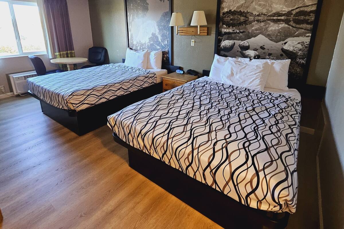 Une chambre d’hôtel avec deux lits, un bureau, une chaise, une lampe et un tableau représentant des montagnes.