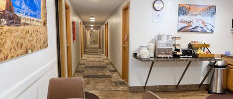 A hotel corridor with a dining area featuring a round table and chairs, a kitchen area with a coffee machine, and a mural on the wall.