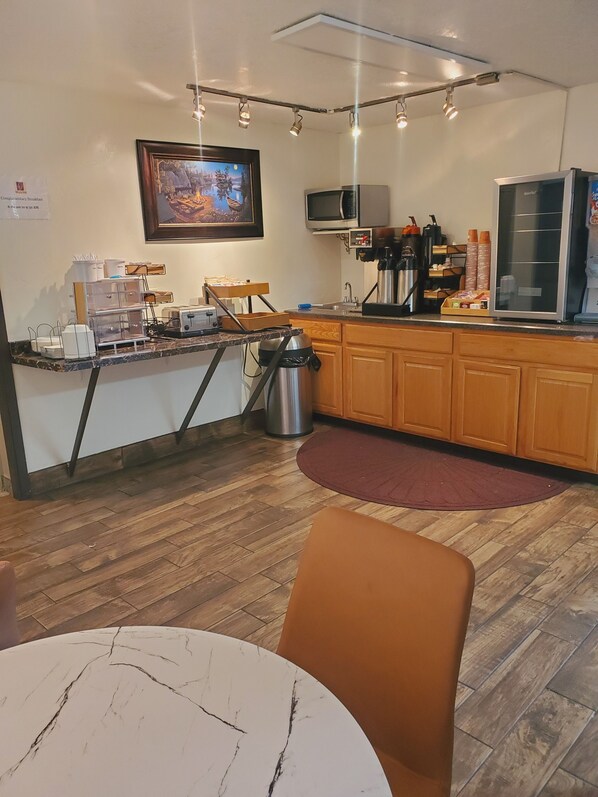A small, well-lit kitchen with wooden cabinets, a microwave, a coffee machine, and a refrigerator. There is a framed picture on the wall and a small table with chairs in the foreground.