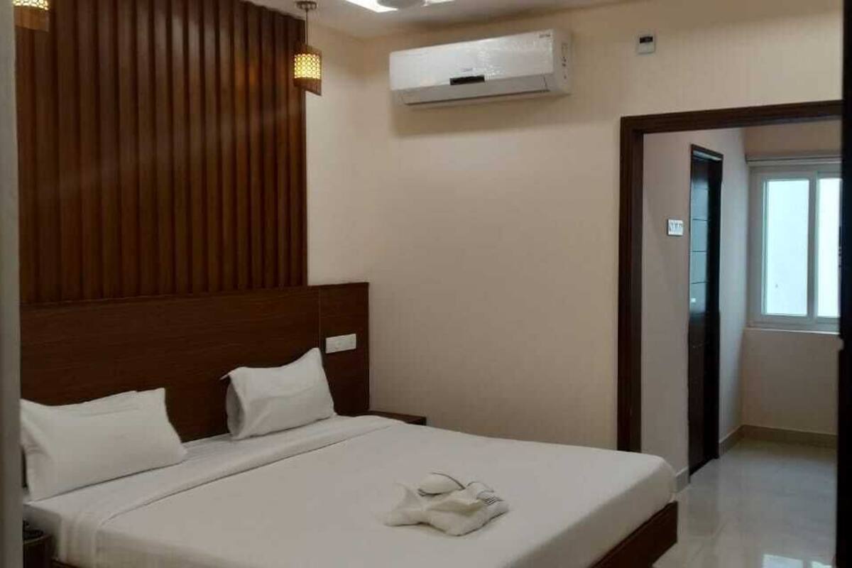 Habitación de hotel con cabecera de madera, cama con ropa blanca, unidad de aire acondicionado y vista al pasillo.