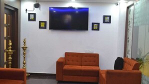 A reception area with a large flat-screen TV mounted on the wall, two orange sofas, a small table with magazines, and a decorative lamp. - AABIL PALACE BUSINESS HOTEL (Chennai)