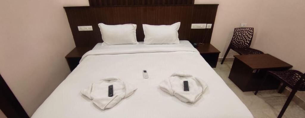 A neatly made bed with two pillows, a bedside table with a remote control, and a chair with a patterned backrest.