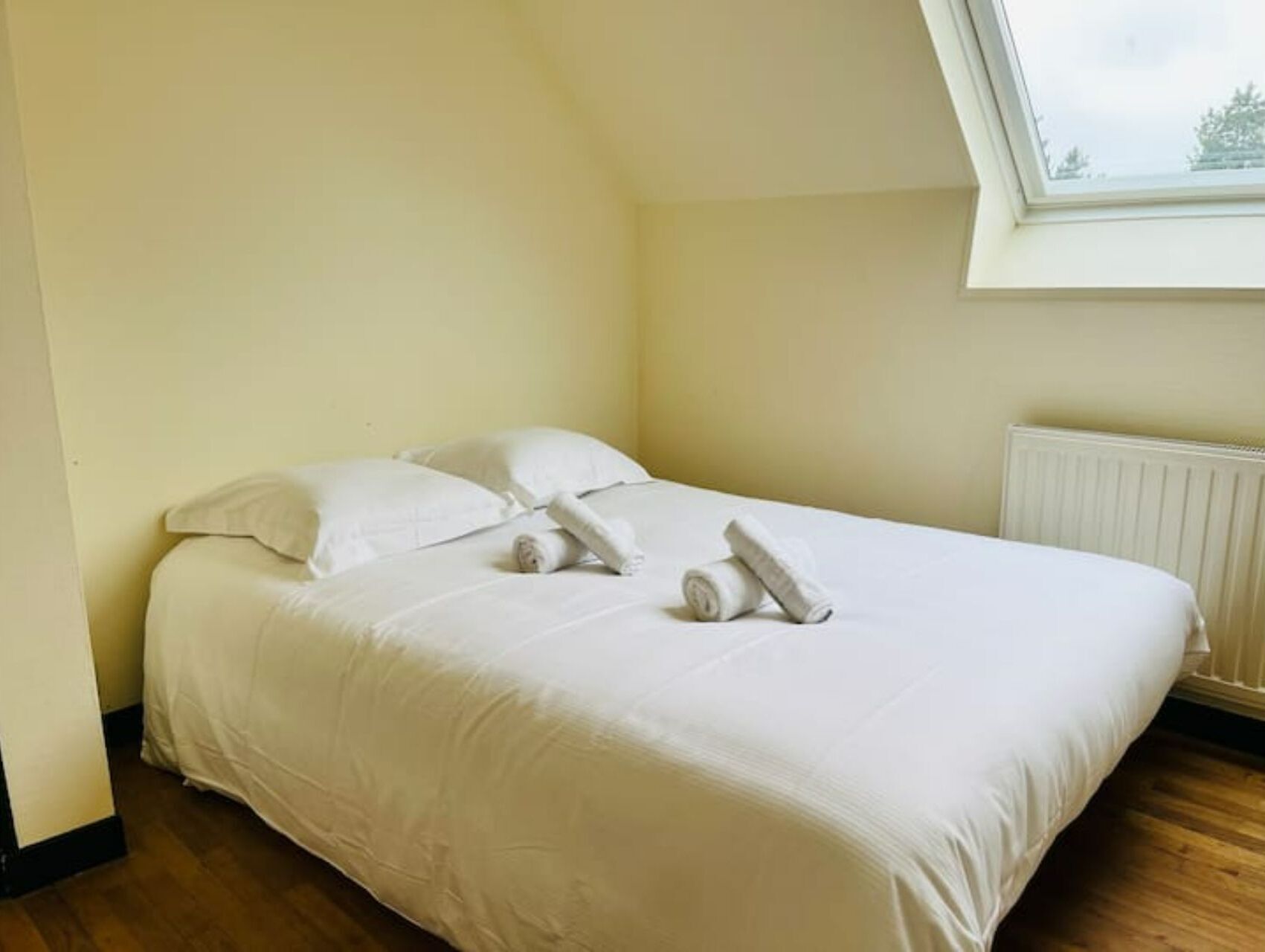 A neatly made bed with white linens and towels, a wooden floor, and a window with a view of trees.