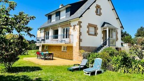 Una casa de dos pisos con un balcón, un patio con muebles de exterior y un jardín con sillones de descanso.