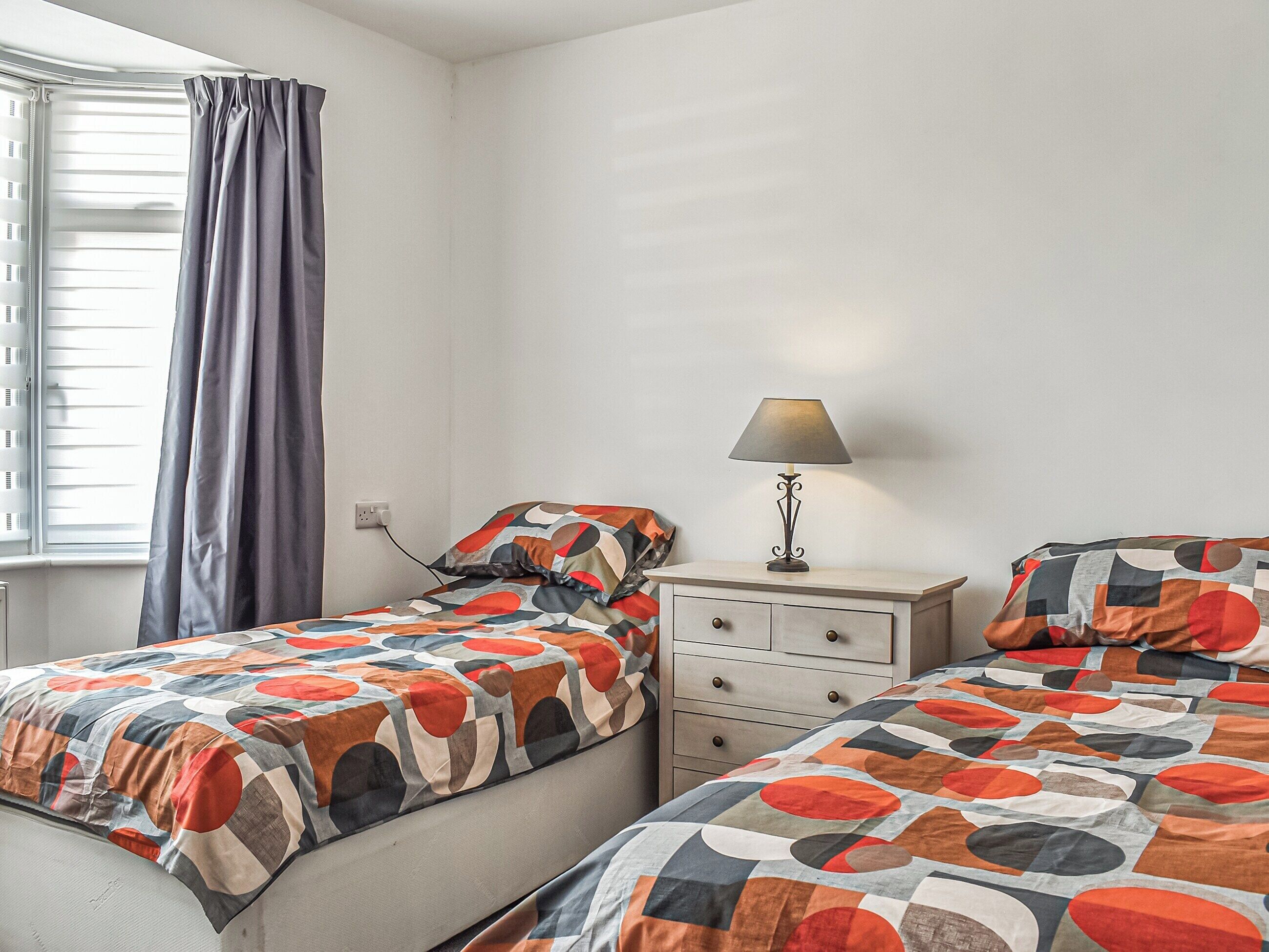 Un dormitorio con dos camas, una mesita de noche, una lámpara y una ventana con cortinas.