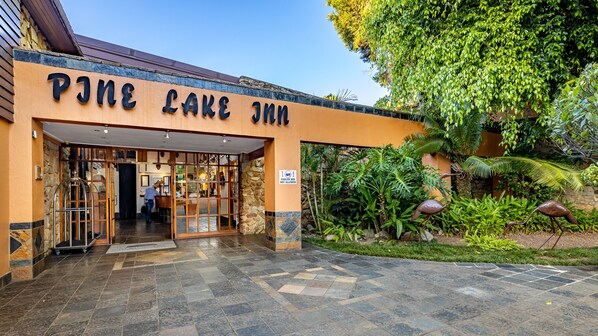 A entrada do P.J. Lake Inn, com fachada de pedra, uma placa indicando desconto de 10% no quarto e um jardim tropical com uma estátua de pássaro.