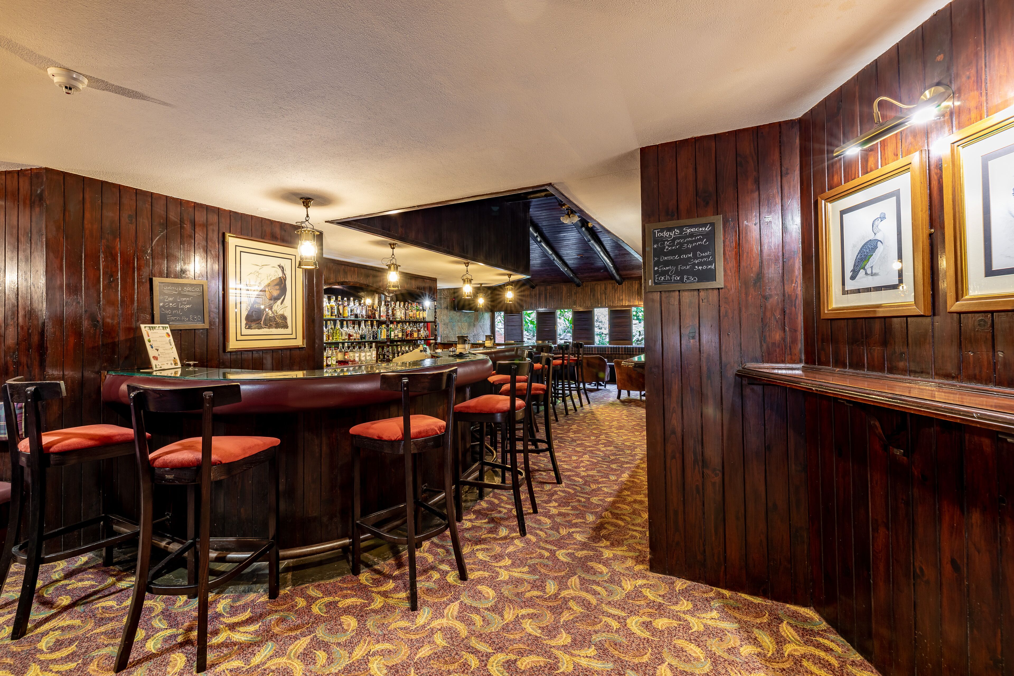 A bar area with wooden paneling, bar stools with red cushions, and framed artwork on the walls.