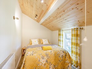 A bedroom with a wooden ceiling, a bed with floral bedding, a bedside table, and a window with curtains.
