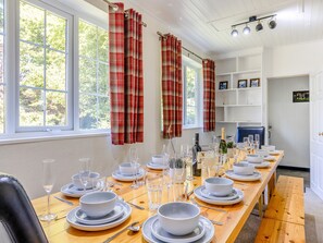 Ein Essbereich mit einem langen Holztisch, der für eine Mahlzeit gedeckt ist, mit Fenstern mit Karottenvorhängen und einem weißen Innenraum mit Regalen und einer Tür.