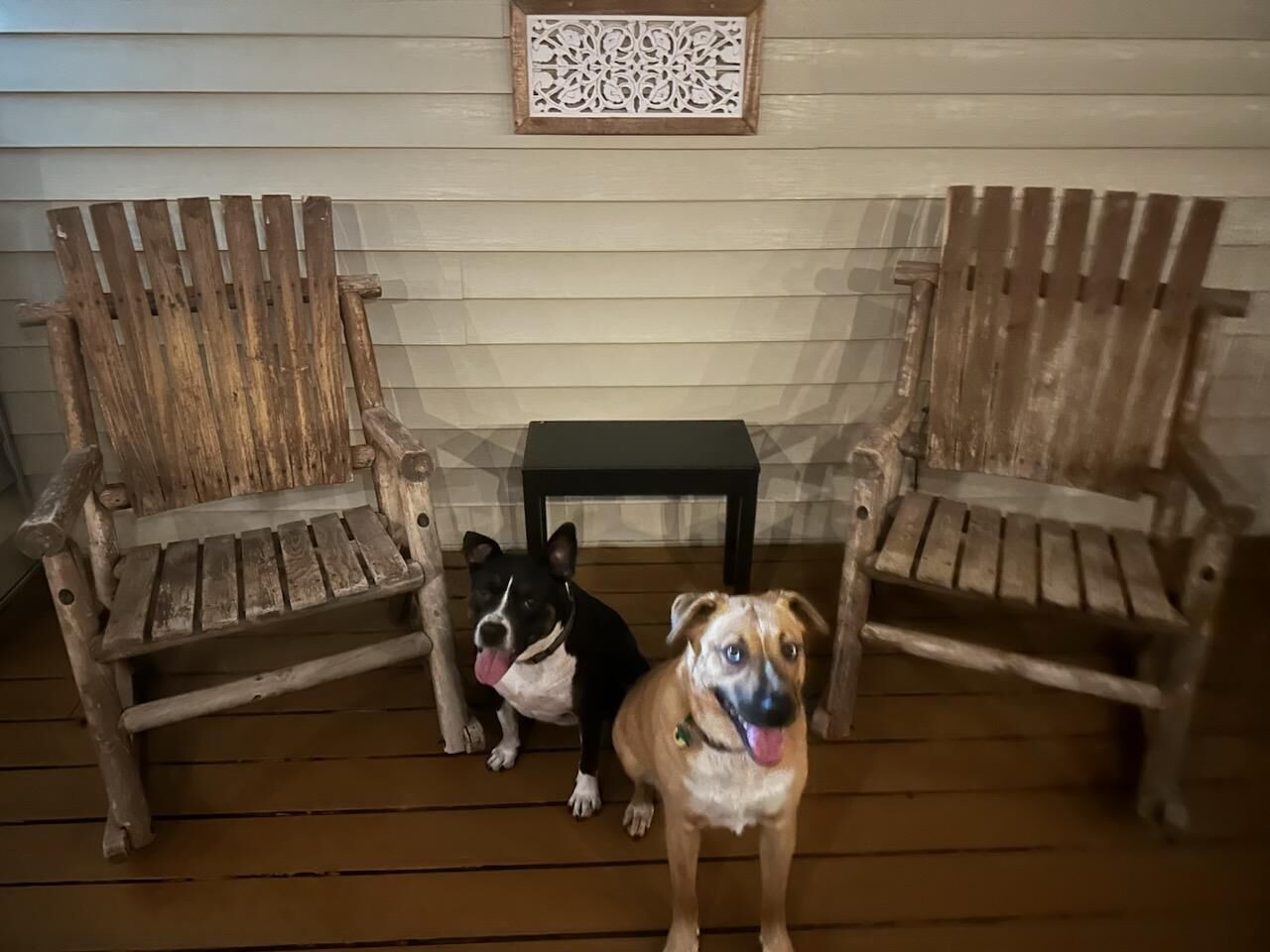 Two wooden chairs and a small table on a wooden deck with a white wall in the background.