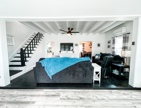 A modern living room with a grey sofa, a staircase, a dining area, and a kitchen visible through an open door. - Coastal Black Point Beach House (East Lyme)