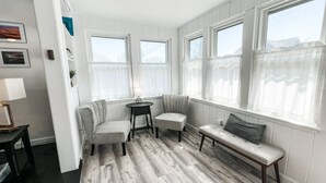 Huoneessa on puulattia, kaksi ikkunaa, joissa on ohuet verhot, penkki ja pieni pöytä, jossa on lamppu.