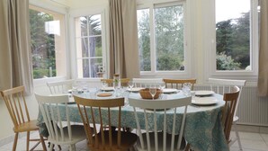 Comedor con una mesa dispuesta para cuatro personas, sillas de madera, ventanas amplias con cortinas y vistas a la vegetación.