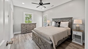 Quarto com cama, mesas de cabeceira, cômoda, ventilador de teto e janela com vista para o verde.