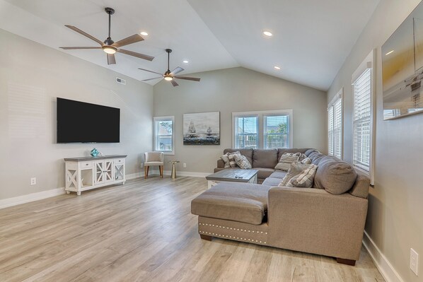 A spacious living room with a large sectional sofa, a flat-screen TV mounted on the wall, a ceiling fan, and a dining area with a small table and chairs.