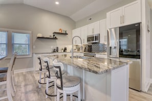 A modern kitchen with white cabinets, a stainless steel refrigerator, a microwave, and a granite countertop. There are two bar stools and a window with blinds.