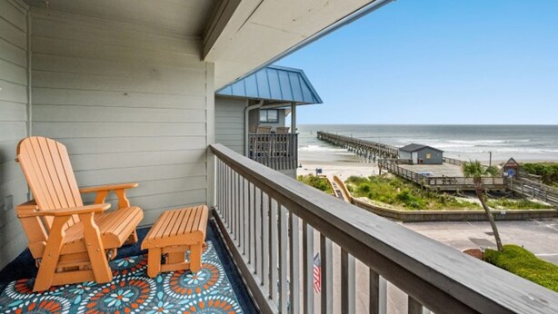 Un balcon avec deux fauteuils Adirondack en bois et une petite table, donnant sur une plage avec une jetée et un poste de surveillance des sauveteurs.
