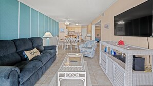 Un salon avec un canapé, une table basse et un téléviseur. On aperçoit à l’arrière-plan un coin repas avec des chaises et une table.