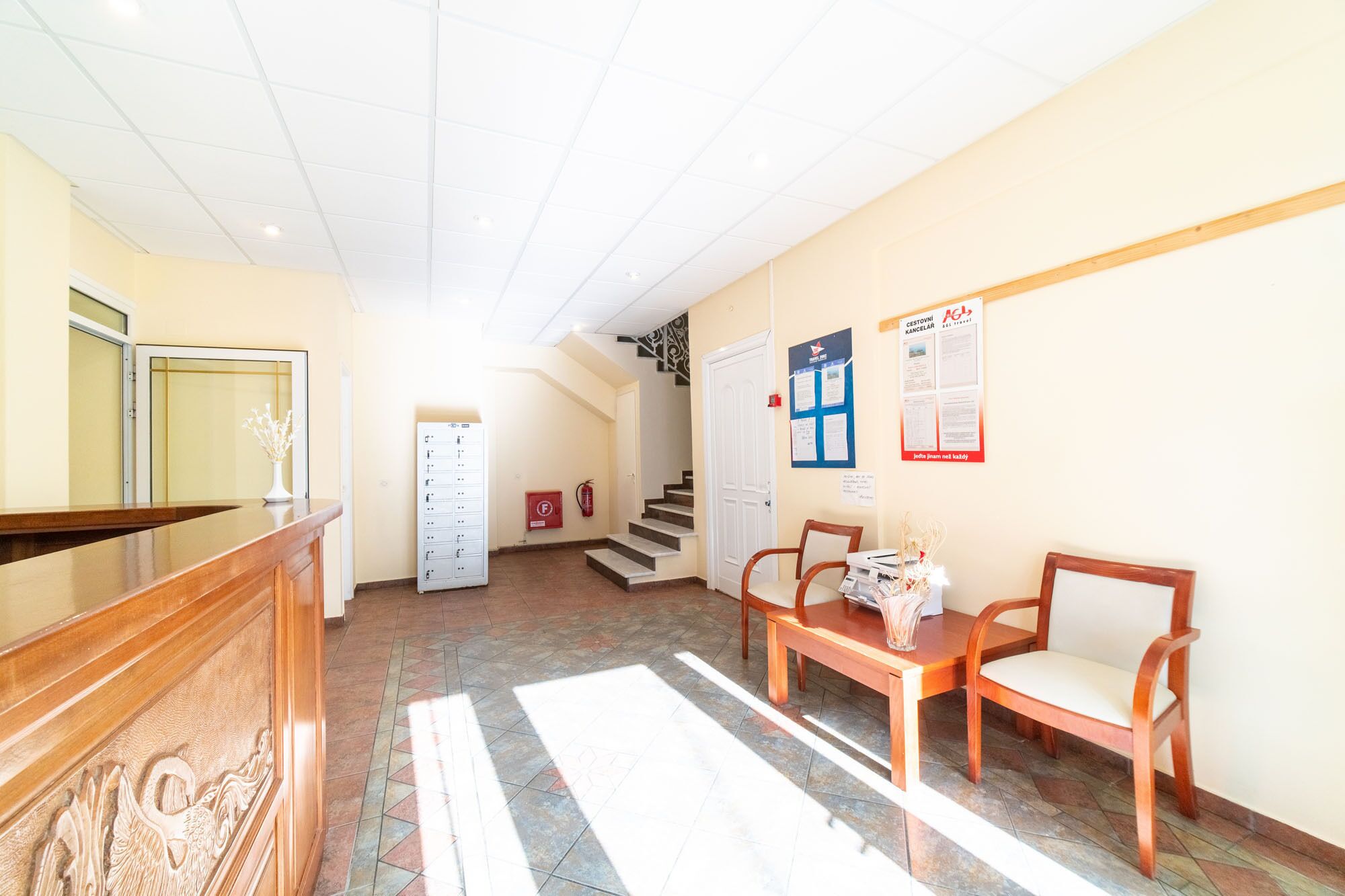 A reception area with a wooden reception desk, a seating area with two chairs, a staircase, and a fire extinguisher on the wall.