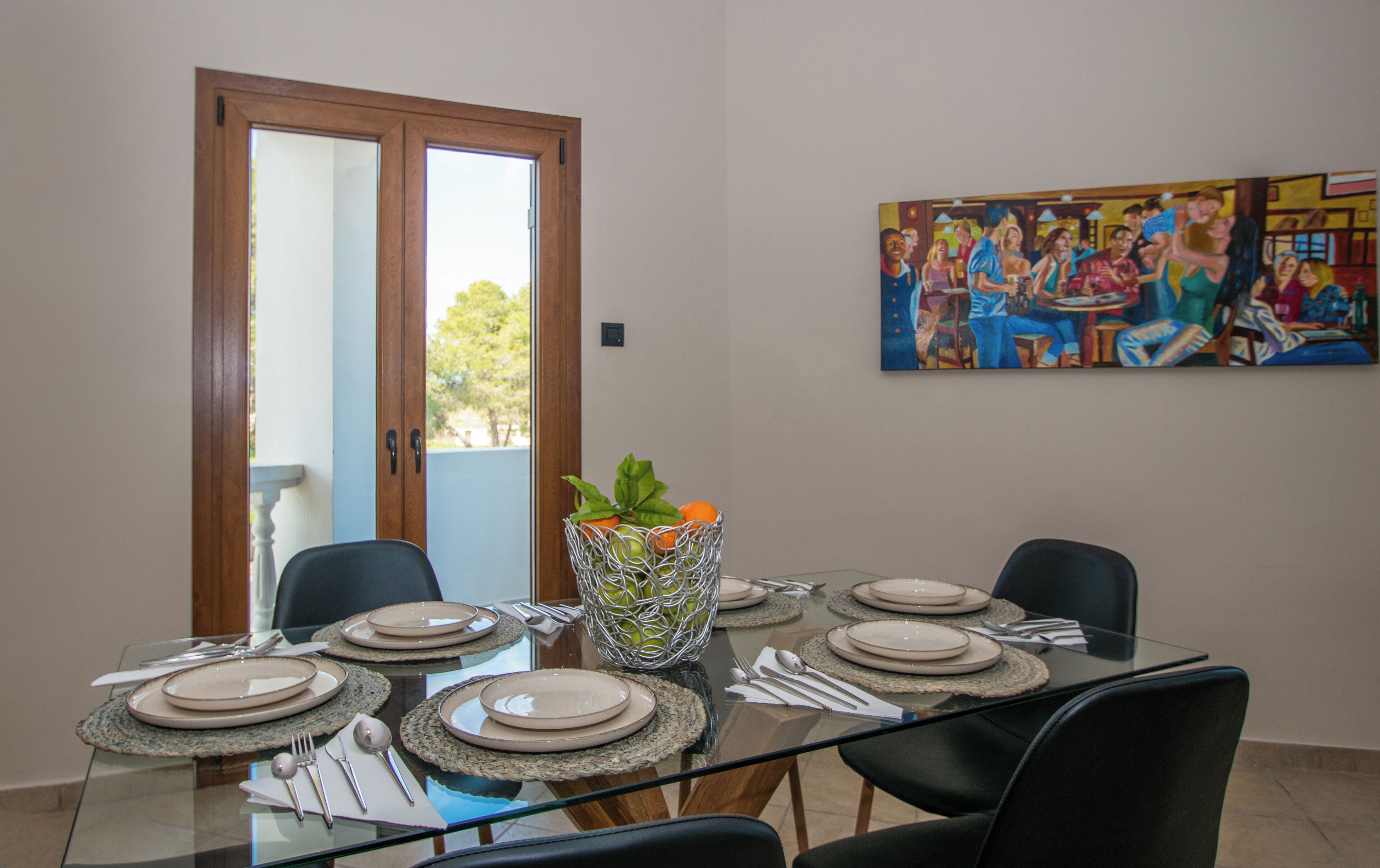 Une salle à manger avec une table en verre dressée pour six personnes, entourée de chaises. Un tableau est accroché au mur, et une porte donne sur l’extérieur.