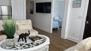 Una sala de estar con un sofá blanco, una mesa de centro de cristal con un libro y una pequeña figurita de elefante, un televisor y una puerta que da a otra habitación.