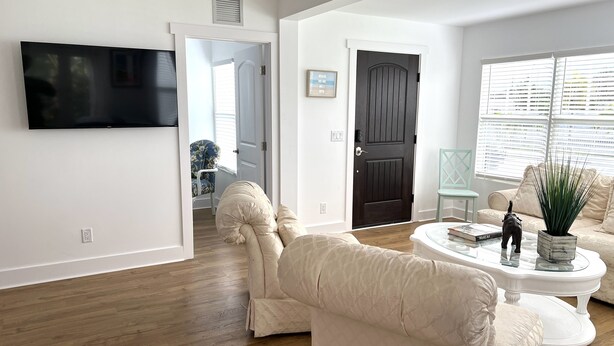 Una sala de estar con una televisión de pantalla plana montada en la pared, una puerta de madera oscura, un sofá color beige y una mesa de centro blanca, además de una planta en maceta.