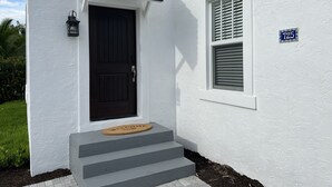 Una casa blanca con puerta negra, una ventana con contraventanas y un felpudo en los escalones.