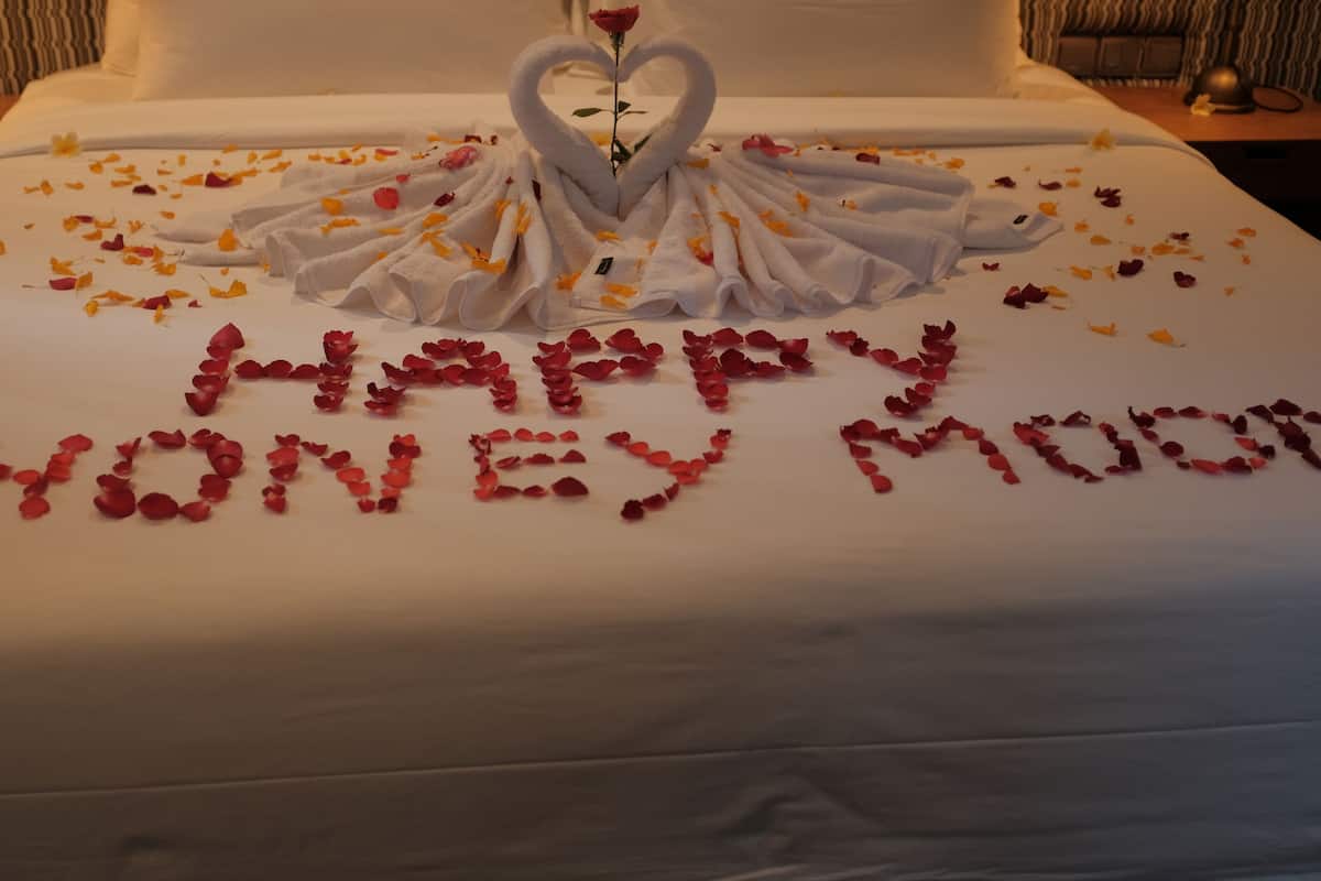 A bed with a heart-shaped towel arrangement and a rose, with 'Happy Honeymoon' written in rose petals.