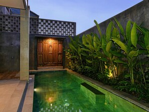 A modern building with a large wooden door, a swimming pool, and lush green plants.