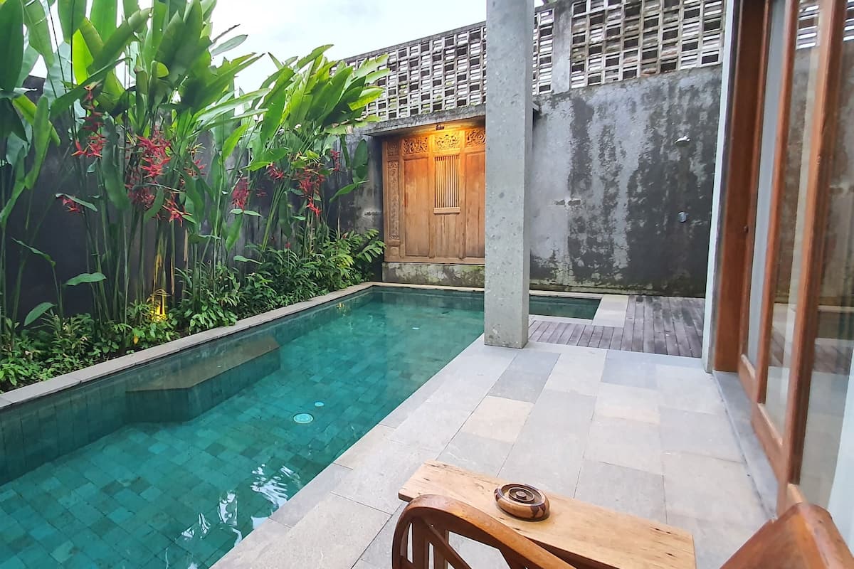 A serene indoor pool area with a wooden table and chairs, surrounded by lush greenery and a traditional wooden door.