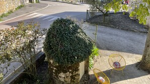 Una strada pavimentata con un muro in pietra e un ingresso ad arco, che conduce a una vista su un lago e montagne. In un'area pavimentata ci sono due sedie gialle con cuscini decorati.