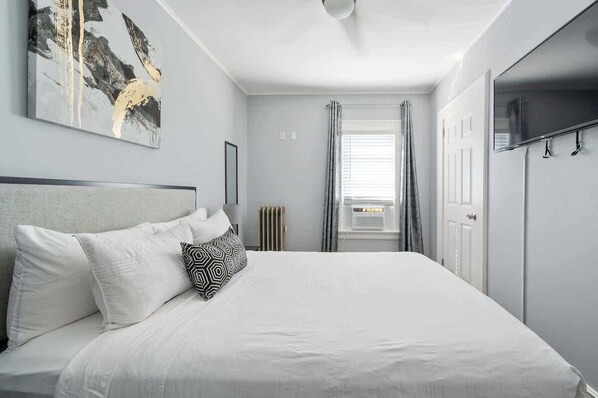 Une chambre à coucher avec un lit, un ventilateur de plafond, une fenêtre avec des rideaux et un téléviseur fixé au mur.