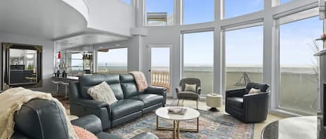 A spacious living room with a large black leather sofa, a round coffee table, and a patterned rug. There are multiple windows offering a view of the sea.