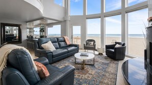 A spacious living room with a large window overlooking a beach, a leather sofa, a coffee table, and a TV. - Luxury Oceanfront Vacation Home on Hollywood Beach (Oxnard)
