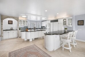 A modern kitchen with white cabinetry, a central island, and a dining area with white wicker chairs. - Luxury Oceanfront Vacation Home on Hollywood Beach (Oxnard)