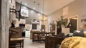 Un salon moderne avec un canapé, une table basse et un téléviseur fixé au mur. On aperçoit à l’arrière-plan un coin cuisine avec une table à manger et des chaises.