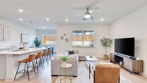 A modern kitchen with a central island, a dining area with a sofa and chairs, a living room with a TV, and a large window with a view of a stone wall and wooden fence. - Spacious Cozy Austin Home with Pool Access (Austin)
