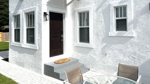 Un edificio blanco con una puerta marrón oscuro, dos ventanas con contraventanas y un pequeño porche con una silla y una mesa.