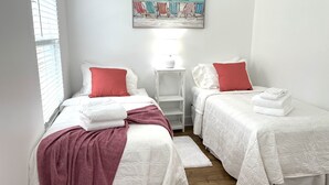 Un dormitorio con dos camas, ropa de cama blanca, almohadas rojas, una manta roja, una mesita de noche blanca, un cuadro con sillas de playa y una ventana con persianas.