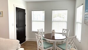 Un área de comedor con una mesa redonda, sillas blancas y un florero. Hay ventanas con persianas, una puerta negra y una pared blanca.