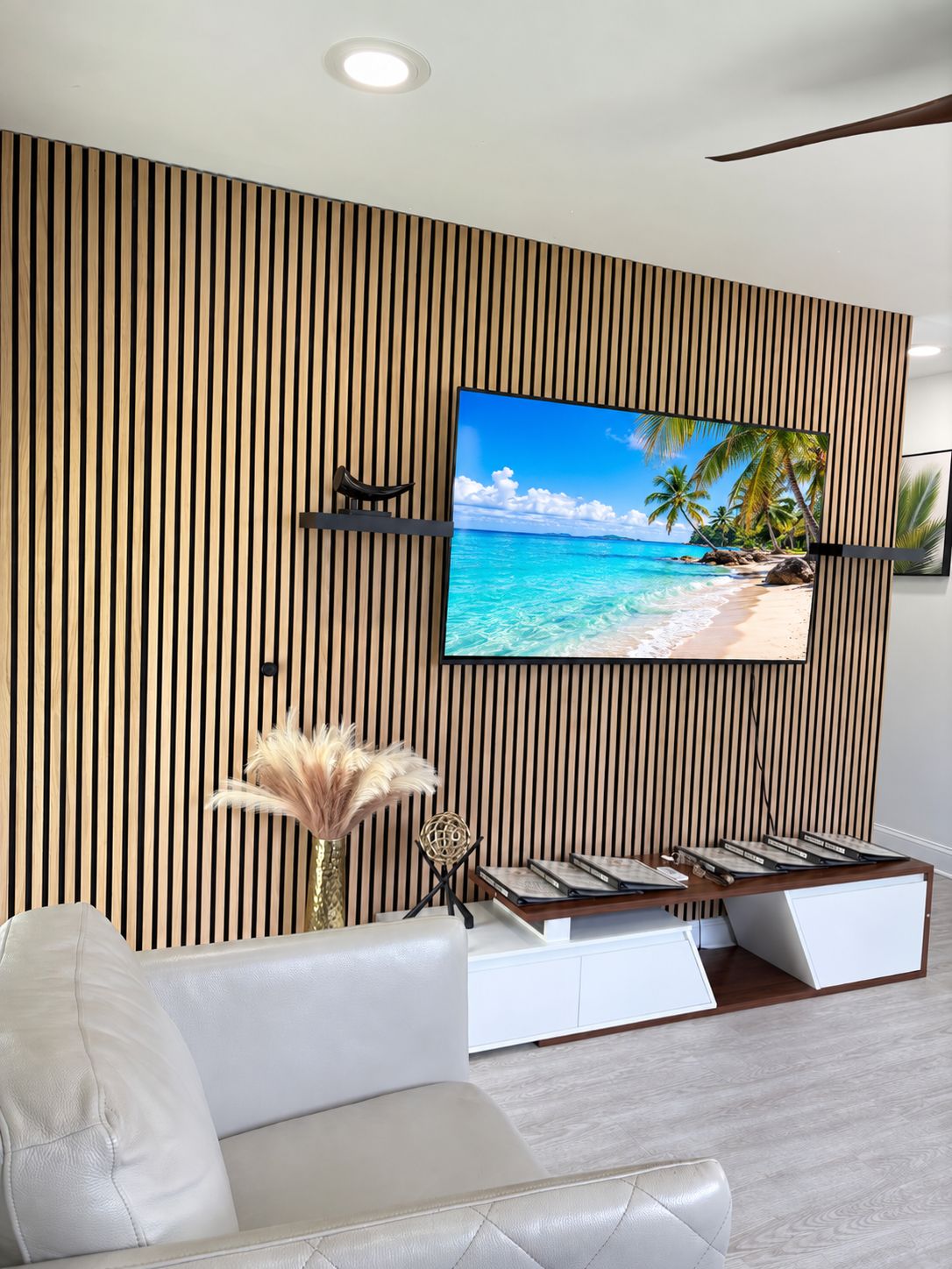 Ett modernt vardagsrum med en stor platt-TV som är monterad på en vägg med träribbor, en vit lädersoffa och ett litet sidobord med dekorativa föremål.
