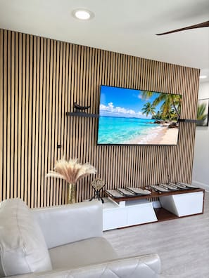 A modern living room with a large flat-screen TV mounted on a wooden slat wall, a white leather sofa, and a small side table with decorative items. - Nova Oasis :- Your private vacation playground (West Palm Beach)