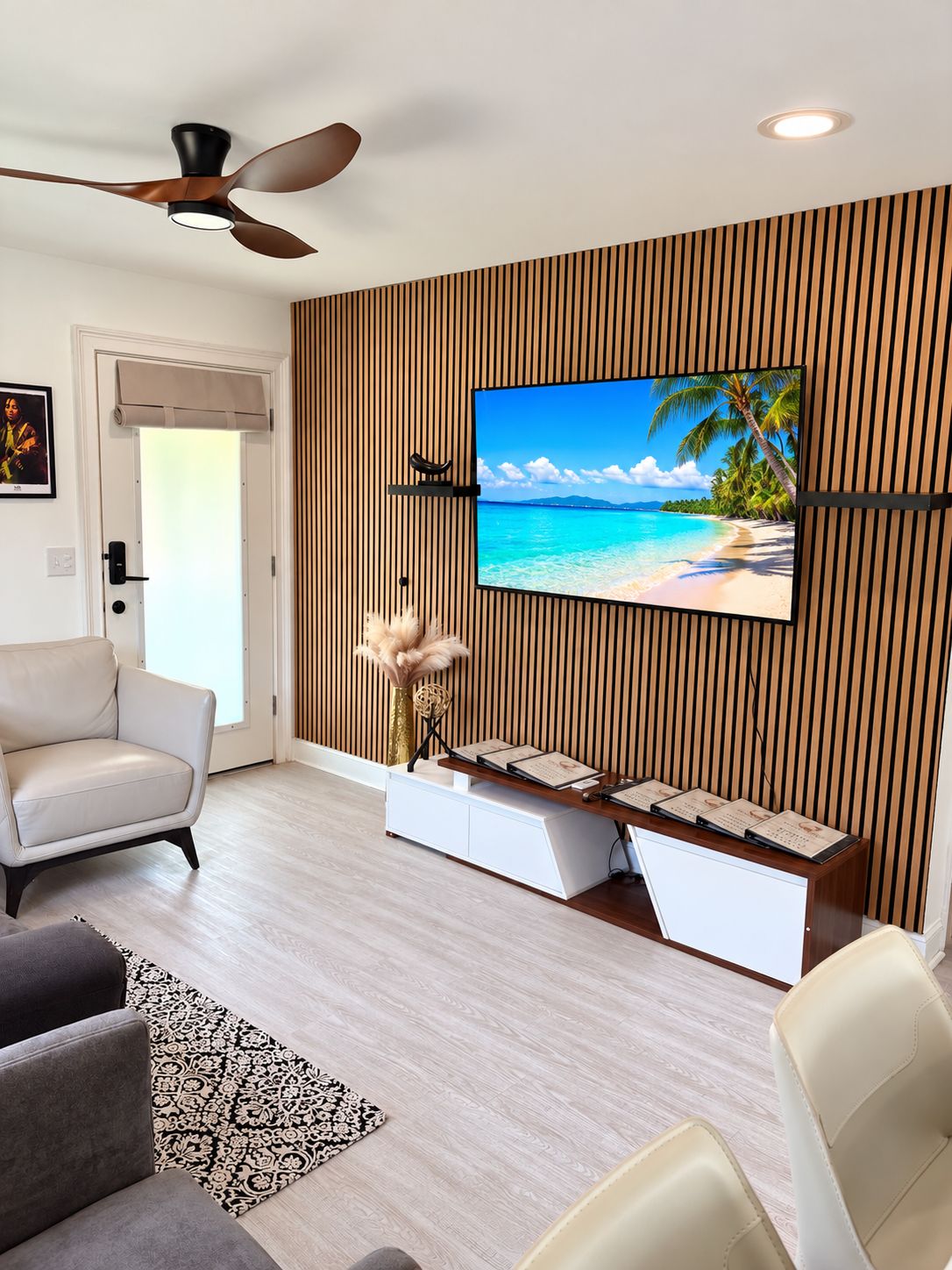 A modern living room with a flat-screen TV mounted on a wooden panel wall, a ceiling fan, and a seating area with a sofa and chairs.