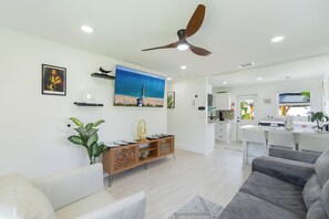 Een moderne woonkamer met een plafondventilator, een grote televisie en een eethoek, met een keuken op de achtergrond.